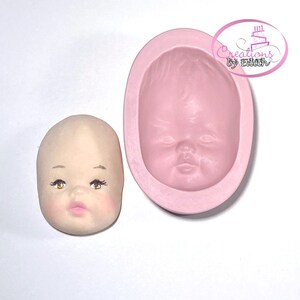 May include: A pink silicone mould for making a baby face cake topper. The mould is shaped like a baby's face with closed eyes, a small nose, and a slightly open mouth. The mould is shown next to a finished cake topper made from the mould.