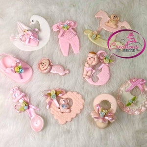 Puede incluir: Un conjunto de 10 favores de baby shower rosas y blancos con una variedad de diseños que incluyen un caballito de balancín, un biberón, un zapato de bebé, un atuendo de bebé, una cuchara, un corazón, un chupete, un babero, un cisne y una muñeca. Los favores están decorados con brillantina y flores.