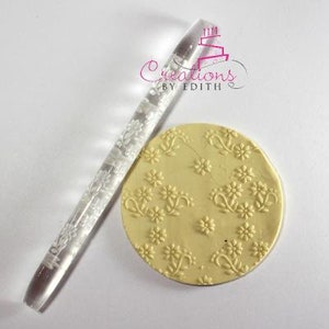 Könnte beinhalten: Ein transparenter Acryl-Nudelholz mit einem Blumenmuster. Das Nudelholz wird verwendet, um dekorative Muster auf Fondant oder Keksteig zu erstellen. Ein Musterbeispiel ist auf einer gelben Fondant-Scheibe zu sehen.