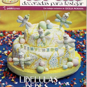 May include: A two-tiered cake decorated with a patchwork design and colourful polka dots. The cake is topped with several fondant dragonflies and pastel-coloured balls. The cake is surrounded by colourful confetti. The text "LIBELULAS BEBES" is printed on the front of the cake. The text "Nacimientos Cumpleaños Bautismos Bodas" is printed below the cake.