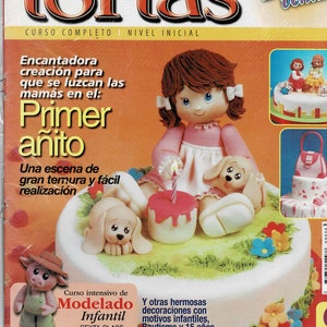 May include: A magazine cover featuring a cake decorated with a doll, two puppies, and a single lit candle. The magazine title is "Decoración de Tortas" and the text "Curso Completo | Nivel Inicial" is below the title. The magazine cover also features the text "Primer Añito" and "Una escena de gran ternura y fácil realización".