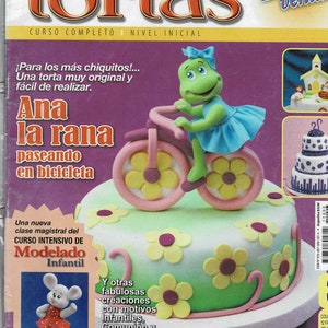 Puede incluir: Una portada de revista con una rana verde montando una bicicleta rosa sobre un pastel decorado con flores amarillas y moradas. El título de la revista es "Decoración de Tortas" y el subtítulo es "Curso Completo | Nivel Inicial".