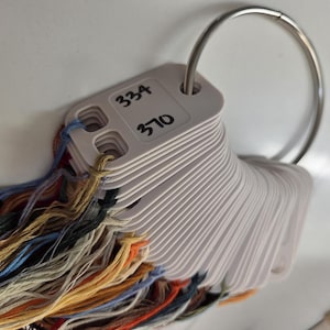 May include: A collection of embroidery floss threads in various colors, including orange, blue, yellow, and grey, attached to white plastic bobbins. The bobbins are organized on a silver metal ring, with the numbers 334 and 370 written on the top bobbin.