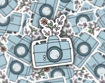 Può includere: Adesivo per fotocamera azzurra con composizione floreale. La fotocamera ha un obiettivo grande e una piccola forma di vaso sulla parte superiore, che contiene alcuni fiori e foglie. Lo sfondo è pieno di adesivi per fotocamere simili.