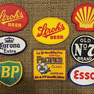 May include: Nine embroidered patches featuring various logos, including Stroh's Beer, Corona Extra, BP, Pacifico Clara, Old No. 7 Brand, and Esso. The patches are all different colors and shapes, and they are all set against a brown background.