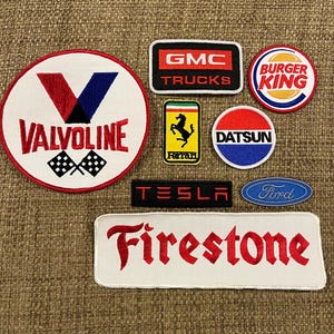 May include: A collection of nine embroidered patches featuring various brands and logos, including Valvoline, GMC Trucks, Burger King, Ferrari, Datsun, Tesla, Ford, and Firestone. The patches are all white with red, blue, and black embroidery.