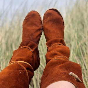 Puede incluir: Un par de botas de ante marr&oacute;n de estilo mocas&iacute;n. Las botas tienen un aspecto suave y texturizado y est&aacute;n atadas con cordones de cuero. Una peque&ntilde;a pieza decorativa de madera est&aacute; unida al lateral de cada bota. Las botas se muestran sobre un fondo de hierba alta.