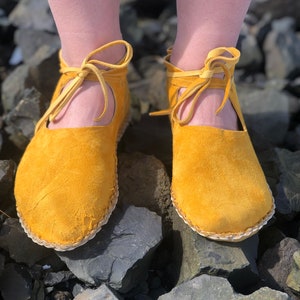May include: Mustard yellow suede leather sandals with ankle ties. The open-sided shoes have a rounded toe and a stitched sole. The sandals are shown on a background of dark gray rocks.