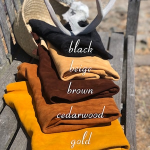 Puede incluir: Cuero de gamuza doblado en negro, beige, marr&oacute;n, cedro y dorado, con etiquetas de color escritas a mano. Una cesta tejida, cornamentas y un banco de madera se encuentran en el fondo. Los colores se muestran en un banco de madera.