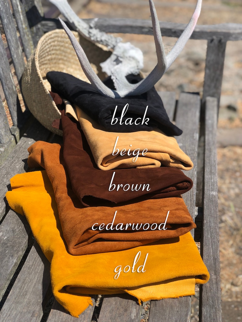 Puede incluir: Cuero de gamuza doblado en negro, beige, marr&oacute;n, cedro y dorado, con etiquetas de color escritas a mano. Una cesta tejida, cornamentas y un banco de madera se encuentran en el fondo. Los colores se muestran en un banco de madera.