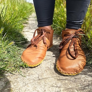 May include: A pair of brown leather moccasin boots with brown laces and yellow stitching around the soles. The boots are worn on a path surrounded by green grass and foliage.