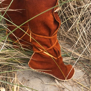 Puede incluir: Botas de ante marr&oacute;n con punta redondeada y parte superior doblada. Las botas est&aacute;n atadas con correas de cuero y presentan un elemento decorativo en forma de cuerno. Las botas se muestran al aire libre, rodeadas de hierba alta.