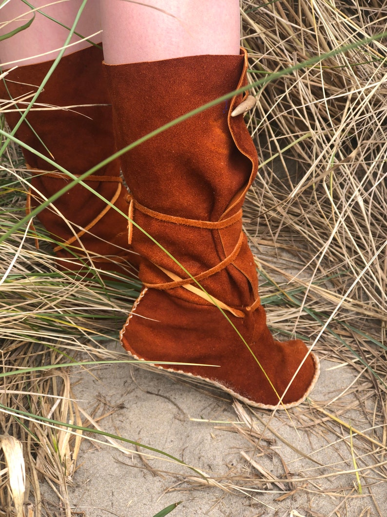 Puede incluir: Botas de ante marr&oacute;n con punta redondeada y parte superior doblada. Las botas est&aacute;n atadas con correas de cuero y presentan un elemento decorativo en forma de cuerno. Las botas se muestran al aire libre, rodeadas de hierba alta.