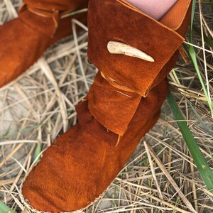 Puede incluir: Primer plano de botas mocas&iacute;n de cuero marr&oacute;n con un pu&ntilde;o doblado y un cierre decorativo de asta. Las botas est&aacute;n hechas de cuero de gamuza suave y est&aacute;n cosidas con un hilo de color claro. Las botas descansan sobre un lecho de hierba seca.