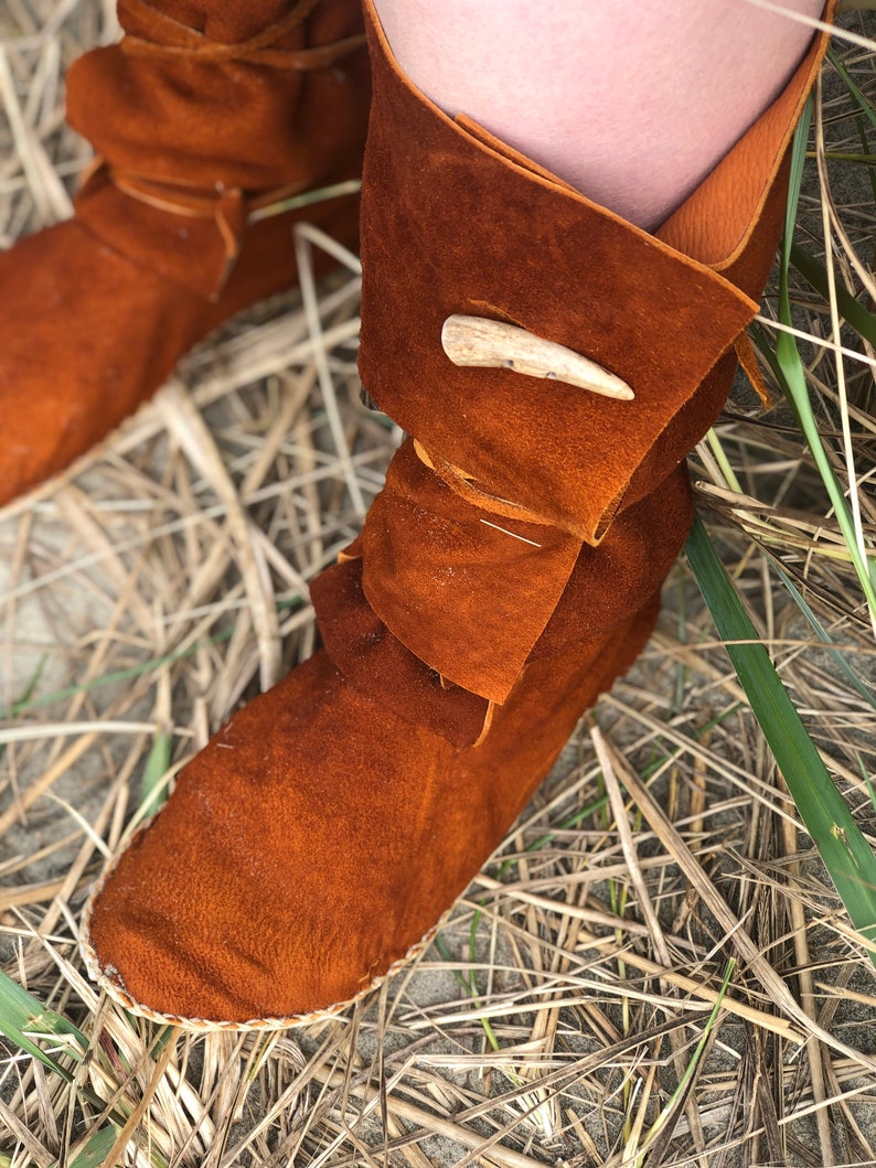 Puede incluir: Primer plano de botas mocas&iacute;n de cuero marr&oacute;n con un pu&ntilde;o doblado y un cierre decorativo de asta. Las botas est&aacute;n hechas de cuero de gamuza suave y est&aacute;n cosidas con un hilo de color claro. Las botas descansan sobre un lecho de hierba seca.