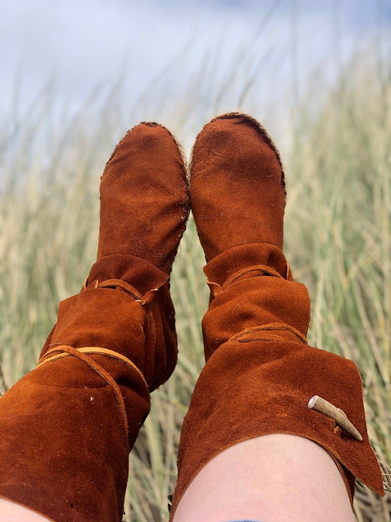 Puede incluir: Un par de botas de ante marr&oacute;n de estilo mocas&iacute;n. Las botas tienen un aspecto suave y texturizado y est&aacute;n atadas con cordones de cuero. Una peque&ntilde;a pieza decorativa de madera est&aacute; unida al lateral de cada bota. Las botas se muestran sobre un fondo de hierba alta.