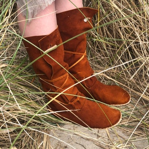 Puede incluir: Botas altas de ante marr&oacute;n con dise&ntilde;o hasta la rodilla. Las botas tienen una parte superior doblada, lazos decorativos de cuero y cierres de bot&oacute;n de madera. Las botas se muestran en un entorno natural con hierba alta.