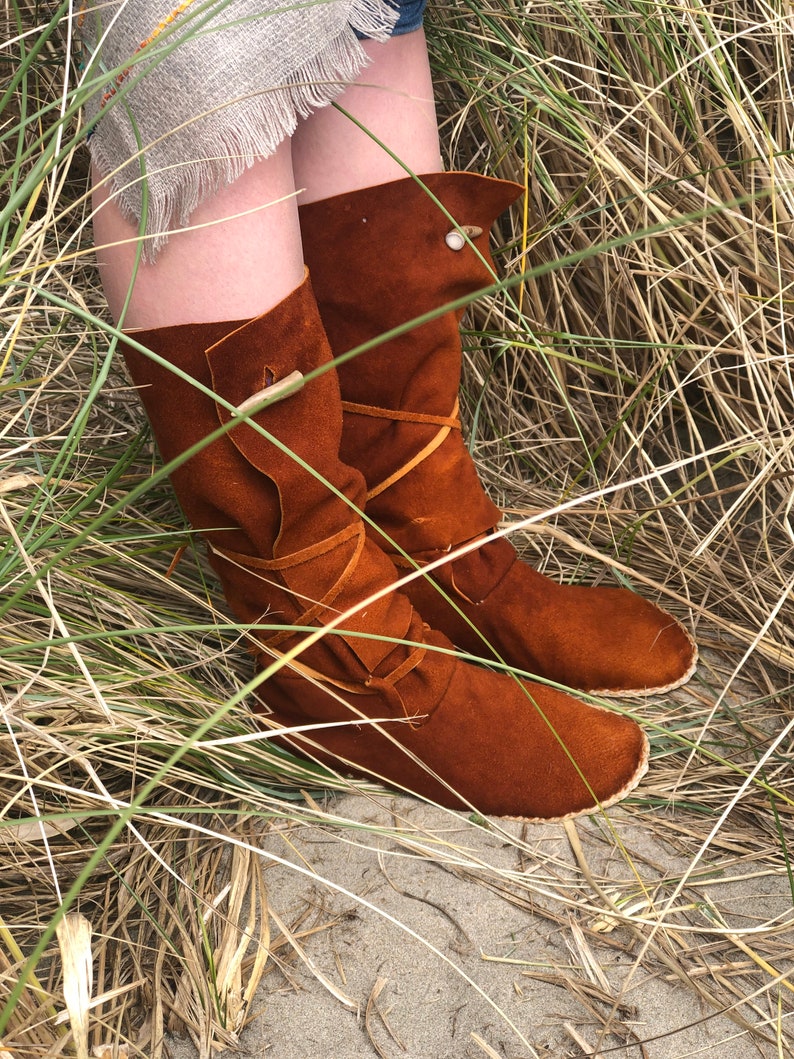 Puede incluir: Botas altas de ante marr&oacute;n con dise&ntilde;o hasta la rodilla. Las botas tienen una parte superior doblada, lazos decorativos de cuero y cierres de bot&oacute;n de madera. Las botas se muestran en un entorno natural con hierba alta.