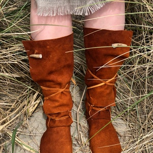 Puede incluir: Botas de ante marr&oacute;n con un dise&ntilde;o r&uacute;stico. Las botas, que llegan hasta la rodilla, tienen una puntera redondeada y una cinta de cuero. Una presilla de madera asegura la parte superior de cada bota. Las botas se muestran en un entorno natural.