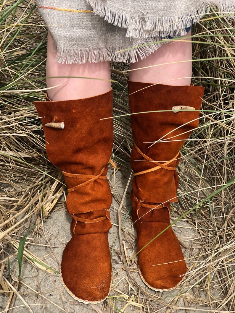 Puede incluir: Botas de ante marr&oacute;n con un dise&ntilde;o r&uacute;stico. Las botas, que llegan hasta la rodilla, tienen una puntera redondeada y una cinta de cuero. Una presilla de madera asegura la parte superior de cada bota. Las botas se muestran en un entorno natural.