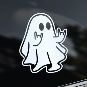 Peut inclure: Un autocollant fantôme blanc aux contours noirs, doté de deux yeux ovales et des deux mains levées dans un geste de rock and roll. L'autocollant est appliqué sur une surface sombre et réfléchissante, avec un petit logo couronne blanc en haut à gauche.