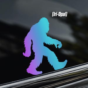 May include: A gradient vinyl decal of a Bigfoot silhouette, transitioning from purple to blue. The text "[Iri-Opal]" is above the figure. A logo with a crown and the letters "CP" is in the upper left corner. The decal is applied to a car window.
