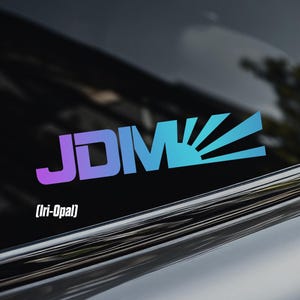 May include: A vinyl decal sticker applied to a car window featuring the text JDM in a gradient color transition from purple to blue, with a stylized sunburst graphic extending from the letter M and the text (Iri-Opal) printed below it.