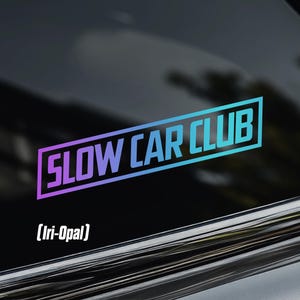 May include: A car window sticker with the text "SLOW CAR CLUB" in a gradient of purple and blue, outlined in a white rectangle. The sticker also includes the text "(Iri-Opal)" below the main text, and a crown logo in the upper left corner.