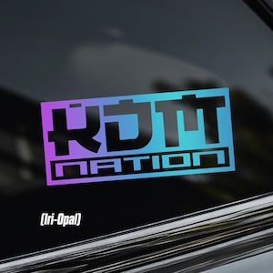 May include: A rectangular sticker with the text "KDM NATION" in a gradient of purple and teal. The sticker is affixed to a car window. The text is in a bold, stylized font. The sticker also has the text "(Iri-Opal)" below the main text.