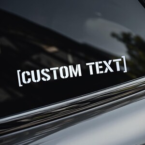 May include: White vinyl decal with the text "[CUSTOM TEXT]" in a stencil font, applied to a car window. The decal is positioned horizontally. A logo of a crown is in the upper left corner.