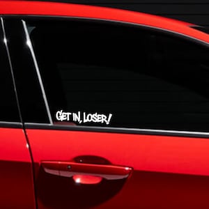 May include: A white vinyl decal applied to the tinted window of a bright red car. The decal features the text "GET IN, LOSER!" in a bold, stylized, sans-serif font. The sticker is positioned on the lower portion of the rear passenger side window.