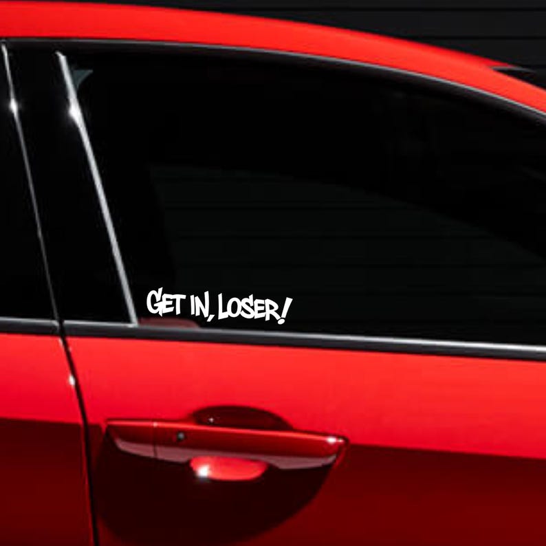 May include: A white vinyl decal applied to the tinted window of a bright red car. The decal features the text "GET IN, LOSER!" in a bold, stylized, sans-serif font. The sticker is positioned on the lower portion of the rear passenger side window.