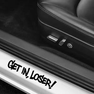 May include: A black vinyl decal applied to the white door sill of a vehicle, featuring the text "GET IN, LOSER!" in a bold, stylized, jagged black font. The decal is positioned below a black leather car seat with visible metal seat controls.