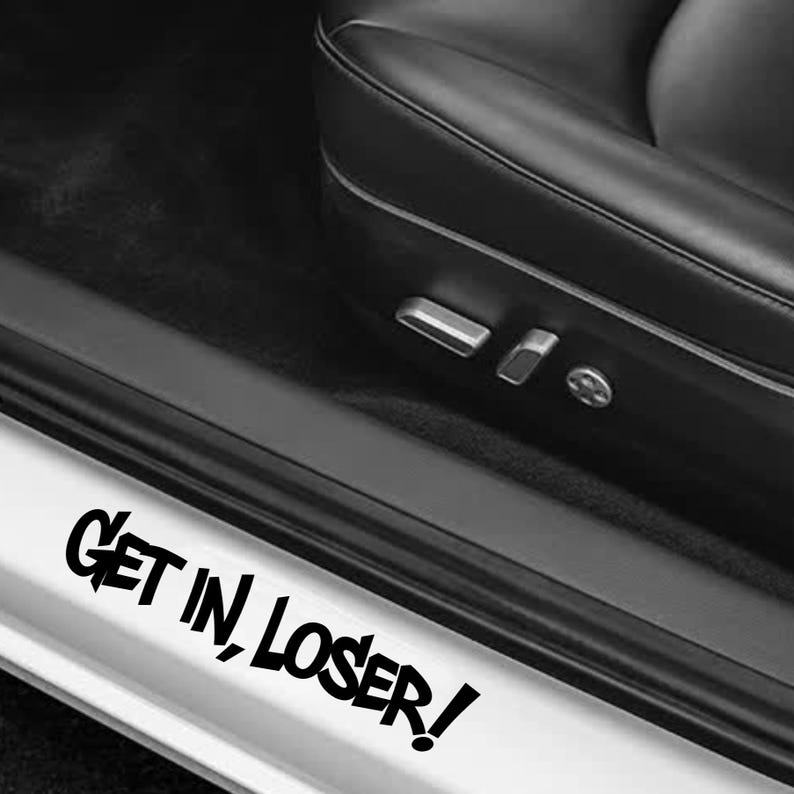 May include: A black vinyl decal applied to the white door sill of a vehicle, featuring the text "GET IN, LOSER!" in a bold, stylized, jagged black font. The decal is positioned below a black leather car seat with visible metal seat controls.