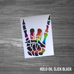 Może przedstawiać: Prostokątna naklejka przedstawiająca dłoń szkieletu wykonującą gest rock and rolla. Wz&oacute;r dłoni ma żywe, opalizujące tęczowe wykończenie na białym tle, z napisem HOLO-OIL SLICK BLACK wydrukowanym poniżej.