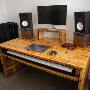May include: A wooden studio desk with a keyboard, computer monitor, and speakers. The desk has a natural wood finish and a built-in keyboard shelf. A laptop and mouse are on the desk. The speakers are black with white speaker cones. A red audio interface sits on the desk.