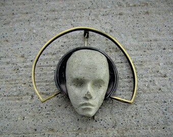 May include: A gray concrete sculpture of a face with a gold metal halo. The sculpture is mounted on a gray concrete wall.