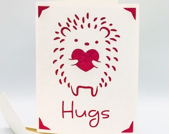 Könnte beinhalten: Eine weiße Karte mit einem roten Igel, der ein rotes Herz hält. Die Karte sagt "Hugs" in rot.