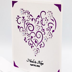 May include: A white card with a purple heart-shaped design featuring intricate swirls and flowers. The text "Nicole & Rhys" and "April 20, 2022" is printed in black at the bottom.