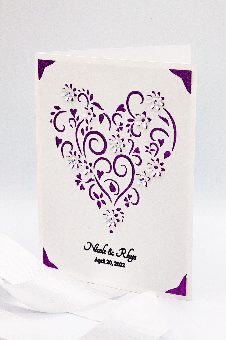 May include: A white card with a purple heart-shaped design featuring intricate swirls and flowers. The text "Nicole & Rhys" and "April 20, 2022" is printed in black at the bottom.