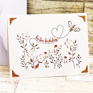 May include: A white card with a brown border and a cut-out design of flowers, leaves, and a bee. The text "ha bee birthday" is written in cursive on the card.