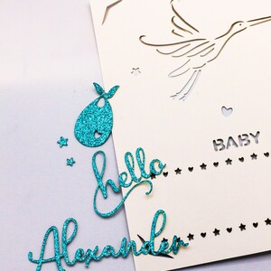 May include: A white card with a cut-out design of a stork flying with a heart and stars. The card has the text "BABY" and a line of stars and hearts. The card is decorated with turquoise glitter paper cutouts of a bib, the words "hello" and "Alexander".