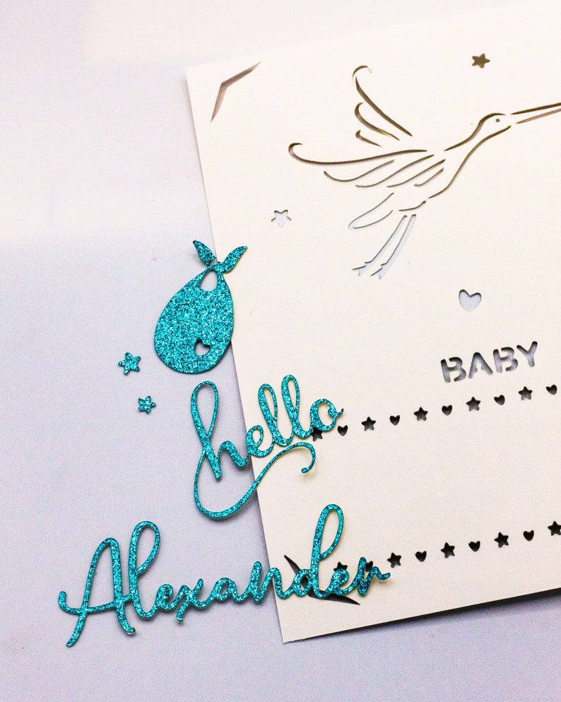 May include: A white card with a cut-out design of a stork flying with a heart and stars. The card has the text "BABY" and a line of stars and hearts. The card is decorated with turquoise glitter paper cutouts of a bib, the words "hello" and "Alexander".