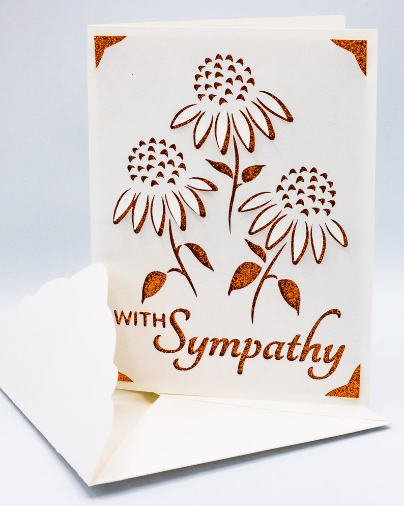 Puede incluir: Una tarjeta blanca con un dise&ntilde;o recortado de tres flores con detalles de purpurina marr&oacute;n. La tarjeta dice "Con simpat&iacute;a" en purpurina marr&oacute;n.