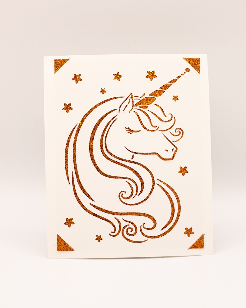 Puede incluir: Una tarjeta blanca con una silueta de unicornio dorada con purpurina y estrellas. El unicornio tiene un ojo cerrado y un cuerno rizado.