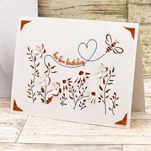 May include: A white card with a brown border and a cut-out design of flowers, leaves, and a bee. The text "ha bee birthday" is written in brown lettering.