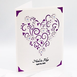 May include: A white card with a purple heart-shaped design featuring intricate swirls and flowers. The heart is decorated with small white flowers and rhinestones. The text "Nicole & Rhys" and "April 20, 2022" is printed in black at the bottom of the card.