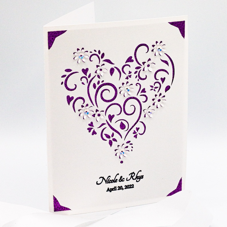 May include: A white card with a purple heart-shaped design featuring intricate swirls and flowers. The heart is decorated with small white flowers and rhinestones. The text "Nicole & Rhys" and "April 20, 2022" is printed in black at the bottom of the card.