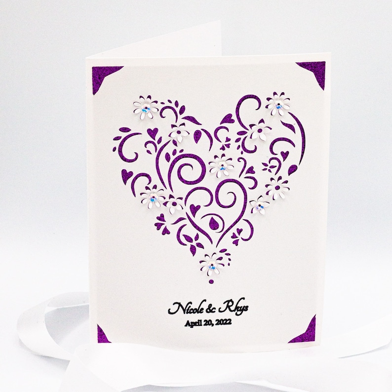 May include: A white card with a purple heart-shaped design featuring intricate swirls and flowers. The heart is decorated with small, clear rhinestones. The text "Nicole & Rhys" and "April 20, 2022" is printed below the heart.