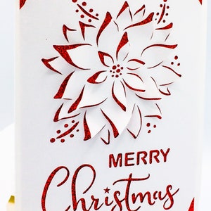 May include: A white card with a red glitter border and a red glitter cutout of a poinsettia flower. The text "Merry Christmas" is written in red glitter below the flower.